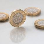 A Sterling pound coin standing on edge on a reflective white surface with three pound coins laying